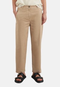 Pantaloni khaki con una vestibilità rilassata, in cotone, due tasche frontali e chiusura con un solo bottone; abbinati a sandali neri con dettagli dorati.