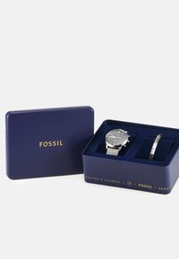 Montre Fossil et ensemble de bracelet dans une boîte en métal bleu marine. La montre présente un cadran gris, un bracelet en maille argentée et des index en chiffres romains. Le bracelet a un design épuré.