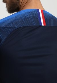 Muž v námořnické modré košili s modrým texturovaným ramenním panelem a vertikálním pruhem v červené, bílé a modré barvě na zadním výstřihu.