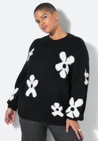 Zwarte gebreide trui met grote witte bloemenpatronen, geribbelde manchetten en een losse pasvorm. Getextureerd materiaal zorgt voor diepte.