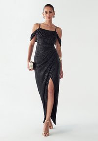 Črna večerna obleka s bleščicami z oprijetim silhuetom, odkrito ramensko obliko, visokim razporkom do stegna in tankimi naramnicami. Zlati dodatki dopolnjujejo videz.
