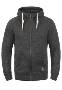 Sudadera gris con cremallera hecha de un tejido texturizado, que cuenta con una capucha con cordón, bolsillos delanteros y un pequeño parche de logo marrón en el lado inferior.