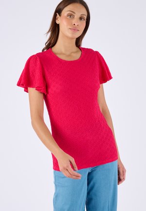 Vrouw draagt een felroze, gestructureerd kortärmelig shirt met vlindermouwen en lichtblauwe spijkerbroek, staand tegen een effen achtergrond.