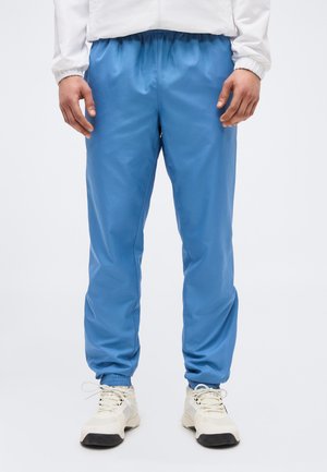 Άνδρας που φοράει μπλε παντελόνι jogger με ελαστική μέση, λευκά αθλητικά παπούτσια και λευκό μακρυμάνικο μπλουζάκι, σε απλό φόντο.