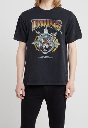 Personne portant un T-shirt noir avec un graphisme de tigre rugissant, des éclairs et le texte "THE KOOPLES" et "HEAVEN ON EARTH."