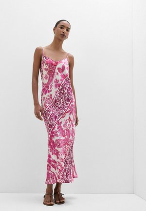 Kvinde iført en hvid og lyserød blomster Paisley-natkjole med tynde stropper og brune sandaler, står mod en ensfarvet hvid baggrund.