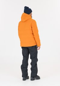 Veste matelassée orange avec capuche, pantalon étanche noir et bottes foncées. La veste présente un design matelassé et des cordons ajustables.