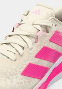 Αθλητικό παπούτσι σε ανοιχτό μπεζ με ροζ λεπτομέρειες. Διαθέτει υφή στην πάνω επιφάνεια, επίπεδα κορδόνια και χαρακτηριστικές ροζ λωρίδες από καουτσούκ στο πλάι.