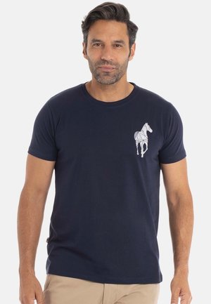 Homme portant un t-shirt bleu marine avec un design géométrique de cheval blanc sur la poitrine gauche, se tenant contre un fond gris clair uni.