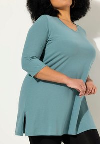 T Shirt tunica a tre quarti di colore teal con scollo a V, spacchi laterali e vestibilità ampia, realizzata in tessuto morbido e leggero.