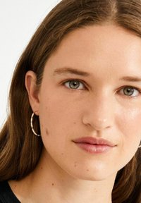 Goldene Creolen mit kleinen weißen Perlen, runde Form, glatte Oberfläche. Das Modell hat mittelbraune Haare und ein natürliches Make-up.