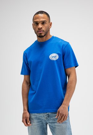 Man draagt een felblauw T-shirt met klein wit logo op de borst en lichtblauwe spijkerbroek, staand tegen een effen lichte achtergrond.
