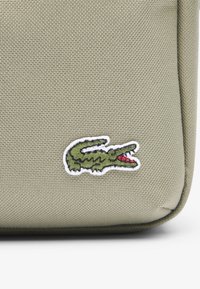 Bolsa de lona verde con una superficie texturizada y un logo de cocodrilo bordado en blanco con acentos rojos.