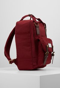 Sac à dos rouge avec poche avant, sangles à boucle en cuir marron et bretelles rembourrées, posé sur un socle blanc devant un fond gris.