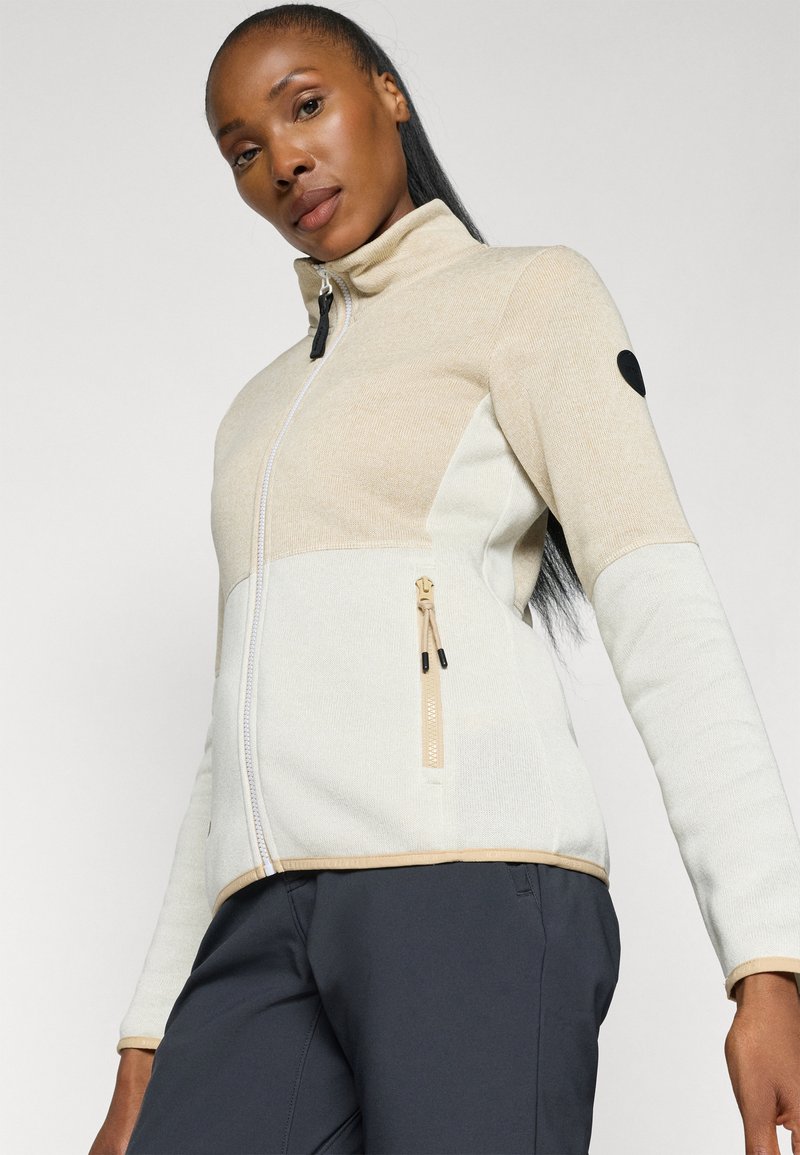 Chaqueta beige y blanca con cremallera y alto cuello, de tejido texturizado y con un bolsillo lateral. Parche de acento negro en la parte superior del brazo.