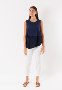 Top senza maniche blu navy con un pannello di un blu navy più scuro nella parte inferiore; abbinato a pantaloni bianchi a sigaretta e sandali a scivolo marroni.