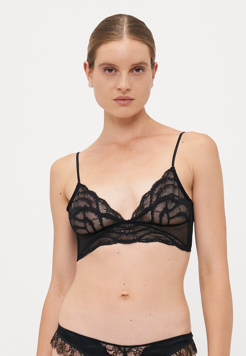 Woman with slicked-back hair wearing a black sheer lace bralette with thin straps and matching lace underwear against a plain background.