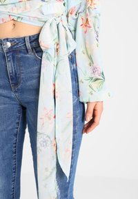 Hedvábná blůza s květinovým vzorem v světle modré barvě s páskem na zavázání, doplněná o tmavě modré seprané džíny, které ukazují různorodé textury a barvy.