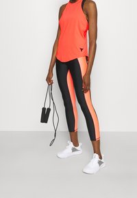 Top senza maniche arancione neón con dettagli neri, abbinato a leggings neri con pannelli laterali arancioni neón e scarpe da ginnastica bianche.