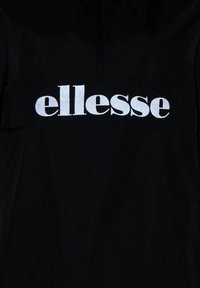 Czarna tkanina z dużym, odblaskowym, białym logo "ellesse" nadrukowanym na obszarze klatki piersiowej.
