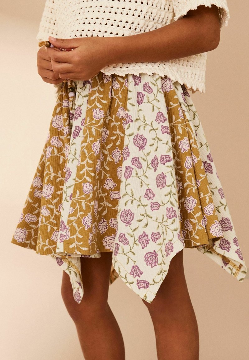 Jeune personne portant une jupe ourlet en foulard à motif floral avec des panneaux alternés moutarde et crème, et un haut blanc en crochet.