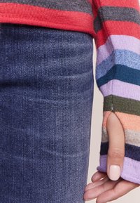 Mehrfarbiger gestreifter Strickpullover in Rot, Grau und Pastelltönen; lange Ärmel mit Daumenlöchern, kombiniert mit dunklen Denim-Jeans.