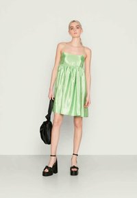 Jovem mulher com um vestido sem mangas de um verde claro brilhante, segurando uma bolsa preta, usando saltos altos pretos, de pé contra um fundo plain.