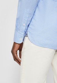 Camicia a quadri azzurra chiara con polsini abbottonati, presenta un orlo curvo. Abbinata a pantaloni color crema, vista dal polso in giù.