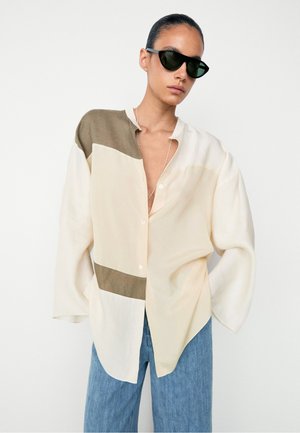 Personne portant une chemise oversize à blocs de couleur beige, crème et marron avec un jean bleu et des lunettes de soleil noires, debout devant un fond blanc uni.