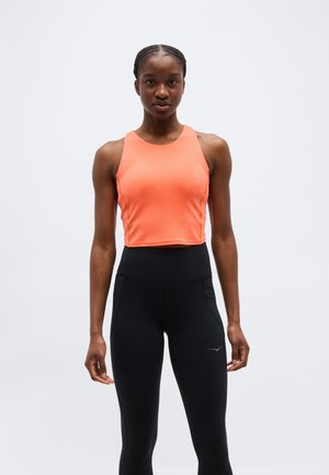 Nő áll, előre néz, narancssárga ujjatlan crop topot és magas derekú fekete sportleggingset visel, egyszínű háttér előtt.