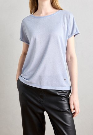 Persoon met een lichtblauw T-shirt met korte mouwen en zwarte leren broek, staand met één hand in een zak tegen een effen achtergrond.
