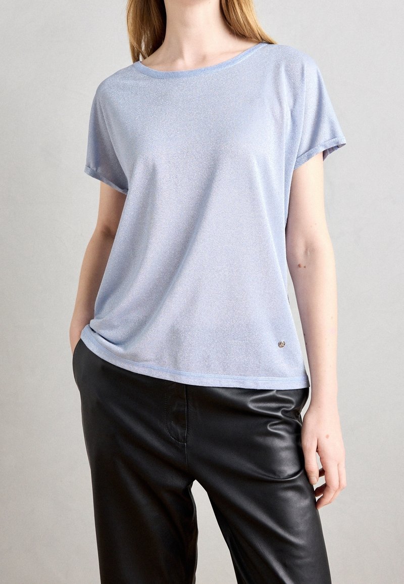 Personne portant un haut bleu clair à manches courtes et un pantalon en cuir noir, debout avec une main dans la poche sur un fond uni.