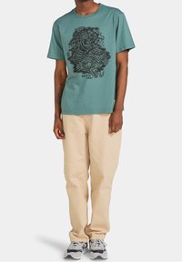 Camiseta de algodón azul verdoso con un diseño gráfico en negro que presenta una figura en medio de olas, combinada con pantalones beige y zapatillas grises.