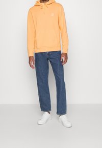 Sweat à capuche orange clair avec poche frontale, associé à un jean droit bleu et des baskets blanches. Design simple, coupe décontractée.