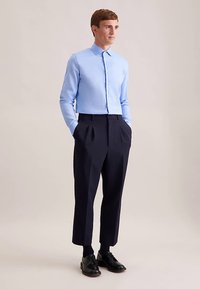 Camicia azzurro chiaro con colletto dritto, abbinata a pantaloni pieghettati blu scuro e scarpe eleganti nere. Tessuto liscio, vestibilità su misura.
