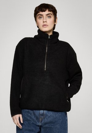 Osoba ubrana w czarną polarową bluzę z półzamkiem i wysokim kołnierzem, w połączeniu z niebieskimi dżinsami, na jednolitym tle.