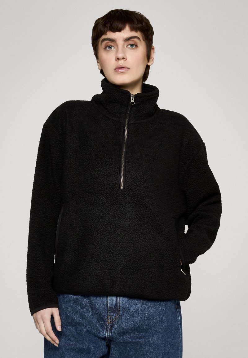 Person iført en sort fleece-pullover med halv lynlås og høj krave, kombineret med blå denimjeans, mod en ensfarvet baggrund.