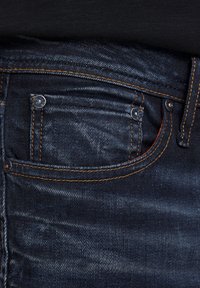 Jeans in denim blu scuro presentano una tasca anteriore con cuciture in rame e hardware a borchia. Il materiale ha una leggera trama e un sottile motivo.