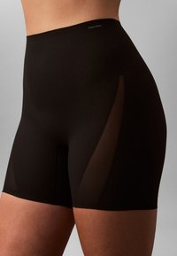 Primo piano di una persona che indossa pantaloncini neri a vita alta con pannelli laterali trasparenti e un piccolo logo Calvin Klein vicino alla vita.