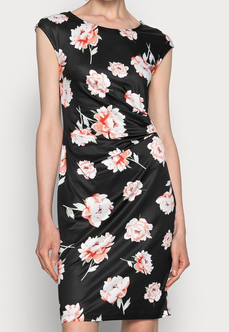 Robe noire sans manches à la silhouette ajustée avec un motif floral rose et blanc, portée par une personne debout devant un fond uni.