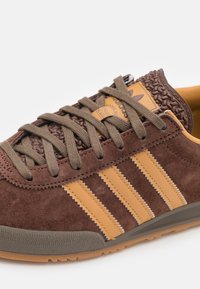 Brown suede sneaker with brown laces, yellow leather stripes, textured brown rubber accents, and a cushioned gray sole.