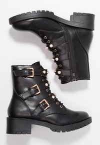 Stivaletti alla caviglia in pelle nera con tacco a blocco, caratterizzati da fibbie dorate, dettagli con perle e una cerniera laterale. Suola in gomma texturizzata per una maggiore aderenza.