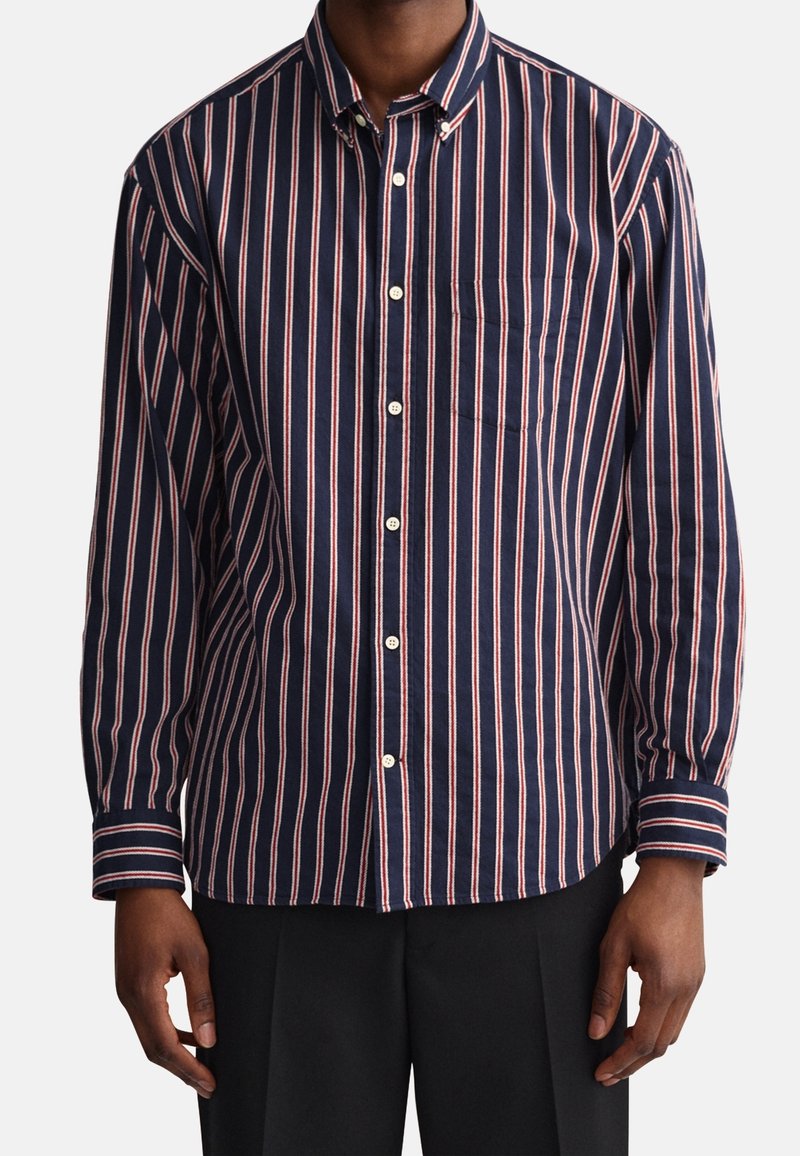 Homme portant une chemise bleu marine à manches longues avec des rayures verticales blanches et rouges et un pantalon noir, debout, les bras détendus le long du corps.