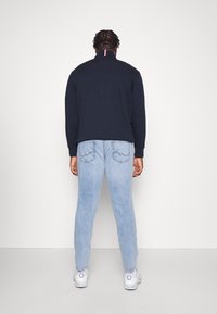 Mörkblå sweatshirts med hög krage och röd/vit/blå detalj på baksidan, kombinerat med ljusblå jeans och vita sneakers.