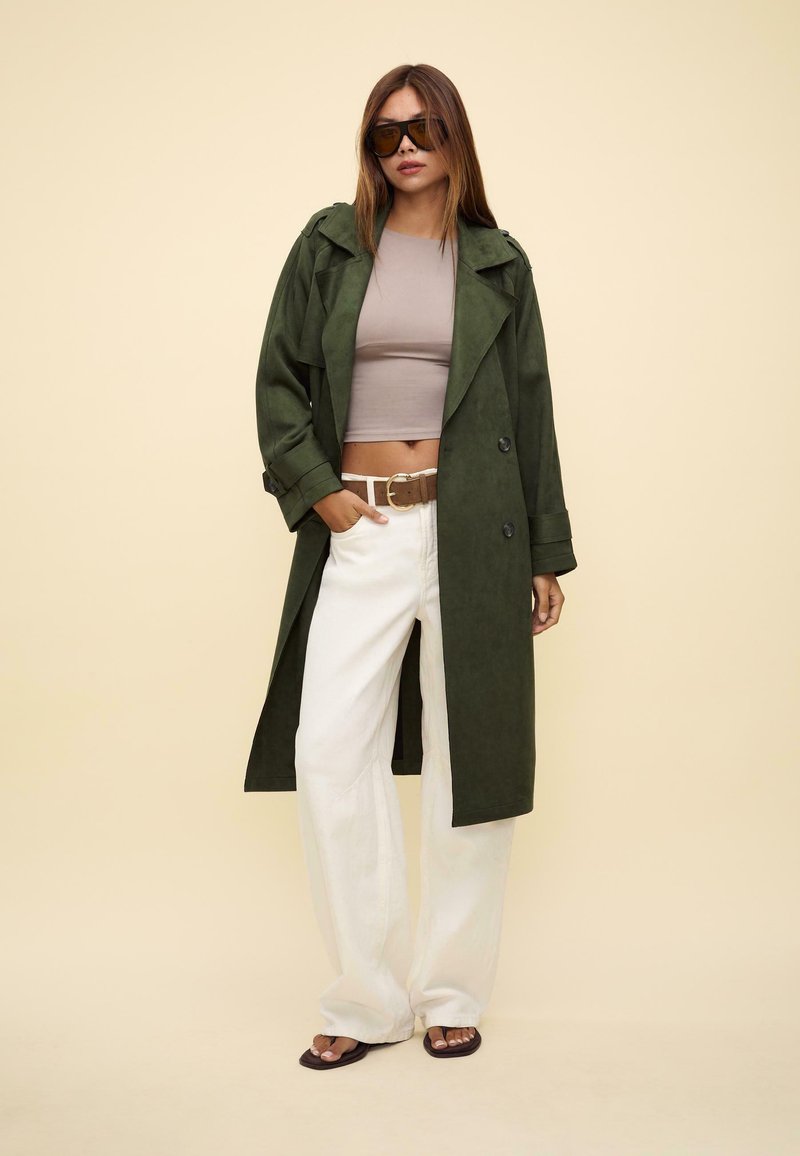 Maslinasto zelena suknja od suede s dvostrukim dugmadi, kremasti crop top, bijele široke hlače i smeđi remen. Tamne sunčane naočale.