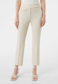 Beige maßgeschneiderte Hose mit hoher Taille, ausgestattet mit zwei Seitentaschen und dekorativen Knöpfen, kombiniert mit weißen Riemchenabsätzen.