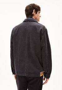 Dunkelgraue Cordjacke mit lockerer Passform, verlängertem Kragen, geknöpften Bündchen und einem beigen Lederlogo-Patch auf der linken unteren Seite.