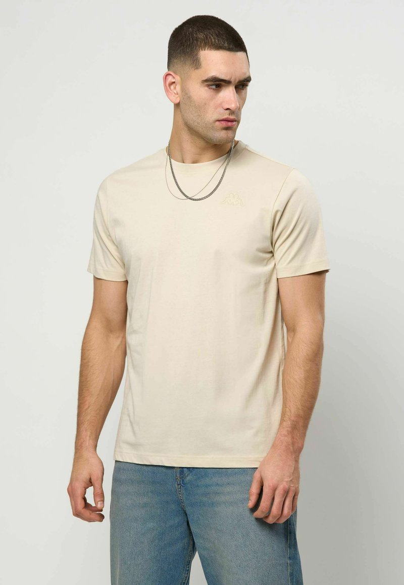 Homme aux cheveux courts et foncés portant un t-shirt beige à manches courtes et un jean bleu clair, regardant vers la droite sur un fond uni.
