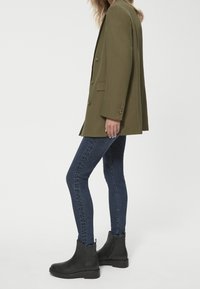 Olive zielona, oversize marynarka z guzikami, zestawiona z ciemnoniebieskimi rurkami i czarnymi botkami za kostkę z teksturowanym detalem.