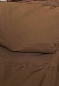 Chaqueta acolchada marrón con un acabado mate, que presenta secciones cosidas y una textura suave. La tela parece ligera y duradera.